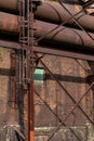 Derelict industrial site with heavily stained brick and rusted metal structure with large tubes, window filled with green corrugat