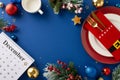 Top view December calendar, plates, whimsical gold flatware in quirky pocket, mug, baubles and more on blue backdrop