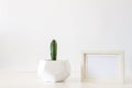 A desk against a white empty wall with a single cactus in a geometric pot and a frame mockup. Royalty Free Stock Photo