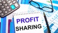 The desktop contains colored tables, a calculator, glasses, a pen and a notebook with the text PROFIT SHARING