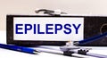 The desktop has a stethoscope, a blue pen, and a gray file folder with the text EPILEPSY. Medical concept