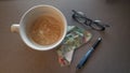 Desktop supplies for work include pen, handkerchief, eye glasses esspresso latte in a white coffee cup ariel view