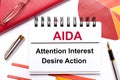 On the desktop is a white notebook with the text AIDA Attention Interest Desire Action, a pen, burgundy and red tables, and gold-