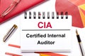 On the desktop is a white notebook with the text CIA Certified Internal Auditor, a pen, burgundy and red tables, and gold-framed