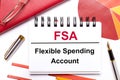 On the desktop is a white notebook with the text FSA Flexible Spending Account, a pen, burgundy and red tables, and gold-framed