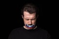 Conceptual portrait of young upset man with three colors duct tape over his mouth isolated on dark background Royalty Free Stock Photo