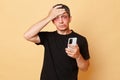 Despair sufferer man in black T-shirt with bruises and abrasions on his face isolated over beige background holding mobile phone