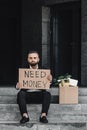 Despaired middle-aged businessman holding cardboard sign with need money text, sitting near business centre, copy space