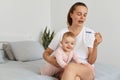 Desperate crying attractive female with pigtail holding pregnancy test in hands, sitting on bed in bedroom with her infant