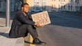 Desperate disgruntled African American millennial student wearing suit sitting on steps outdoors holding poster need