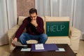 Desperate young man calculating household expenses sitting on the couch and asking for help on a blackboard