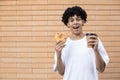 Smiling African-American guy holding a cup of coffee and an orange frosted donut, looking at the camera Royalty Free Stock Photo