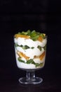 Dessert layered in a transparent glass, yogurt, orange, kiwi, nuts on a dark background. The concept of healthy, proper and simple