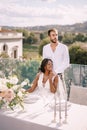 Destination fine-art wedding in Florence, Italy. African-American bride sits at a wedding table, Caucasian groom hugs