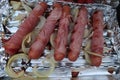 Detail of grilled sausages in ham roll with onion around, placed on disposable aluminium grill plate.