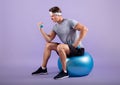 Determined millennial sportsman exercising with dumbbells while sitting on fitness ball on lilac studio background