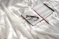 Diary, pencil and eyeglasses rest on white bedding in sunlight with shadows. Personal journal for notes.