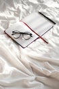 Diary, pencil and eyeglasses rest on white bedding in sunlight with shadows. Personal journal for notes.