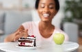 Diet breakdown concept. Young black lady reaching for cake instead of apple, choosing junk food over healthy one at home