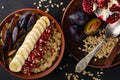 Diet breakfast of a bowl with oat porridge, banana, pomegranate seeds and plum on black background. Top view, flat lay