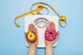 Diet concept. Female hands hold sweet donut and red apple against the background of scales and a measuring tape, choice between Royalty Free Stock Photo