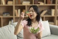 Diet concept. Young korean lady eating healthy food, holding bowl of fresh salad, sitting on sofa at home, free space Royalty Free Stock Photo