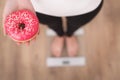 Diet Concept. Young Woman Measuring Body Weight On Weighing Scale While Holding Glazed Donut With Sprinkles. Sweets Are Unhealthy Royalty Free Stock Photo