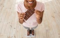 Diet failure, cheat meal and unhealthy nutrition. Above view of black woman holding chocolate on scales, closeup