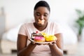 Diet restrictions. Curvy black woman with adhesive bandage on her mouth looking at plate of sweets in hunger at home Royalty Free Stock Photo