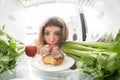 Diet struggle: A girl sadly looking at a donut inside a fridge full of healthy stuff.