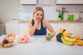 Diet struggle. Young sad woman in blue T-shirt choosing between fresh fruit vegetables or sweets in the kitchen. Choice