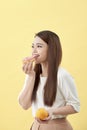 Dieting concept. Beautiful Young Asian Woman holding orange and donut over yellow background. Choosing between junk food with Royalty Free Stock Photo