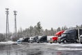 Different big rig professional semi trucks with semi trailers standing at row on the winter truck stop parking lot with snow and Royalty Free Stock Photo