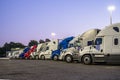 Different big rigs semi trucks standing in row on the night truck stop parking lot with turned on lanterns on poles waiting for