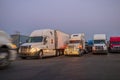 Different big rigs semi trucks with semi trailers standing in row on the twilight truck stop and illuminated by the light of