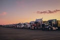 Different make big rigs semi trucks tractors with loaded semi trailers standing in the row on truck stop parking lot at early