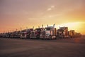 Different make big rigs semi trucks tractors with loaded semi trailers standing in the row on truck stop parking lot at early