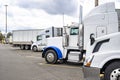 Different make and models of white big rig semi trucks with semi trailers standing in row on truck stop parking lot for truck Royalty Free Stock Photo