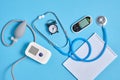 Digital blood pressure monitor, glucometer, stethoscope, notepad and alarm clock on blue background top view