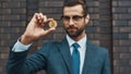 Digital currency. Handsome businessman in formal wear and eyeglasses holding bitcoin in one hand and looking at it while Royalty Free Stock Photo