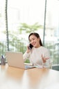 Digital nomad. Young female entrepreneur talking on mobile phone, looking at laptop and smiling, happy woman working in cafe or Royalty Free Stock Photo