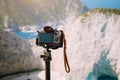 Digital photo camera on tripod against huge cliff rocks of Navagio beach in morning sun light. Famous visiting landmark Royalty Free Stock Photo