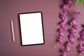 Digital tablet with white screen, stylus pen and flowers on purple background.