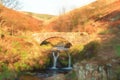 Digital watercolour of an autumnal waterfall and stone packhorse bridge at Three Shires Head in the Peak District.