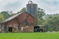 Dilapidated Red Barn in a Green Pasture next to an old Rusty Barn Silo surrounded by a wooded forest in Southern Indiana America` Royalty Free Stock Photo