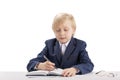 Diligent student writes in copybook. Child like boss sits at table and writes in notebook. Blond boy in jacket and shirt