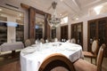 Dining room of a restaurant with oval tables with tablecloths and crockery and wooden upholstered chairs and a pendant lamp in the