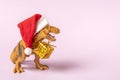 Dinosaur Rex in red Santa Claus hat holds golden gift box in its paws on pink background New Years Eve or Christmas Eve Art Royalty Free Stock Photo