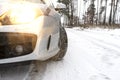 The dipped headlights on a silver car standing on a snowy road in the forest, visible new winter tire and falling snow.