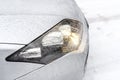 The dipped headlights on a silver car standing on a snowy road in the forest, visible snow falling.